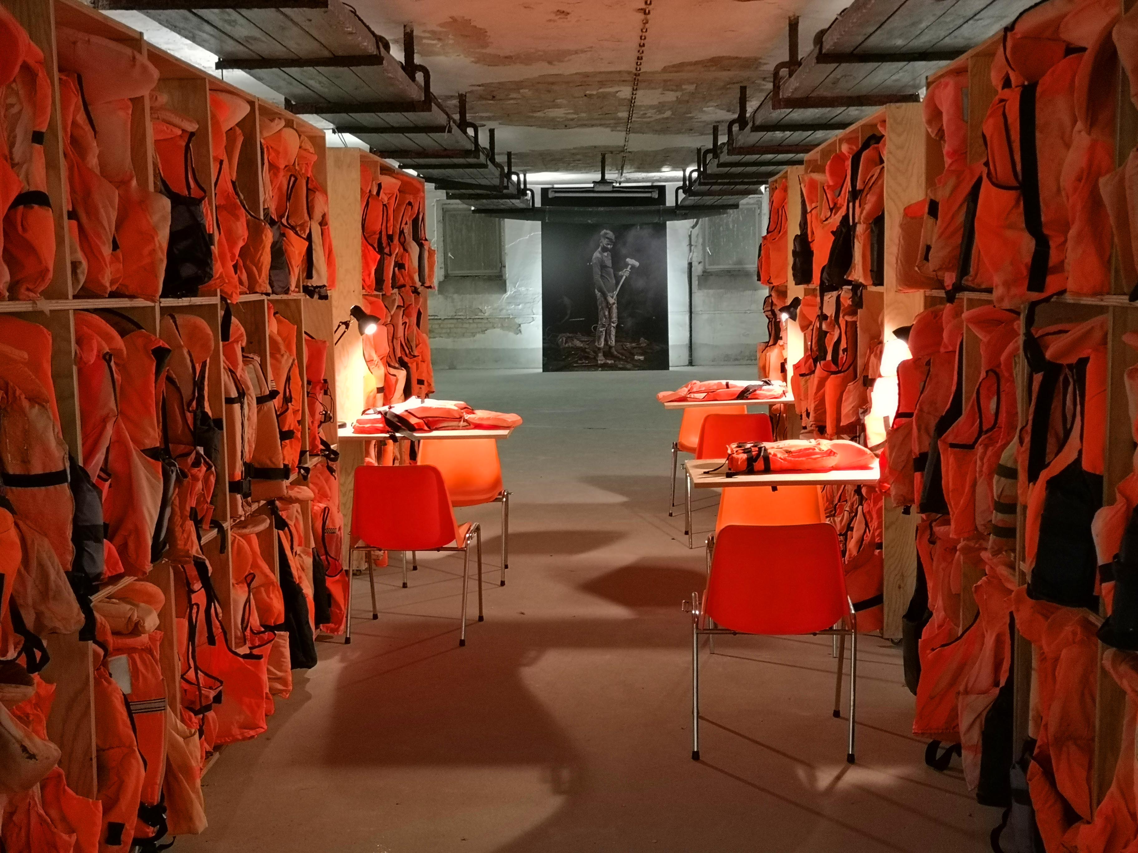 Rauminstallation mit orangefarbenen Schwimmwesten in hohen Regalfächern und roten Stühlen.