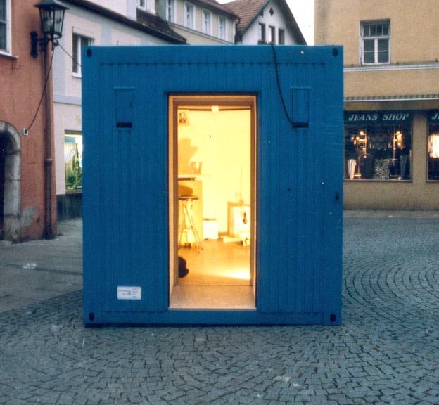 Beleuchteter Eingang des blauen Containers auf einem Platz bei Dämmerung.