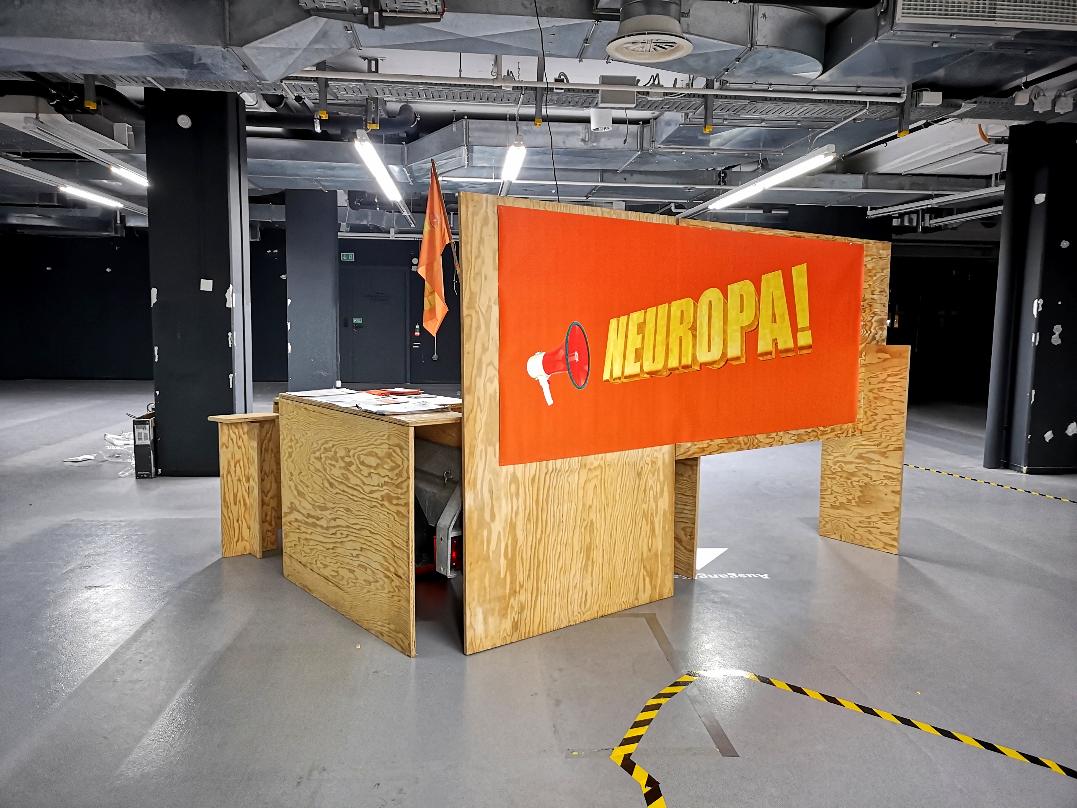 Holzkonstruktion mit dem orangefarbenen Schild „NEUROPA!“ in einem Ausstellungsraum; der Boden ist schwarz-gelb markiert.