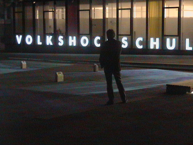 Eine Person steht bei Nacht auf einem Platz; im Hintergrund leuchtet die Aufschrift „VOLKSHOCHSCHULE“.