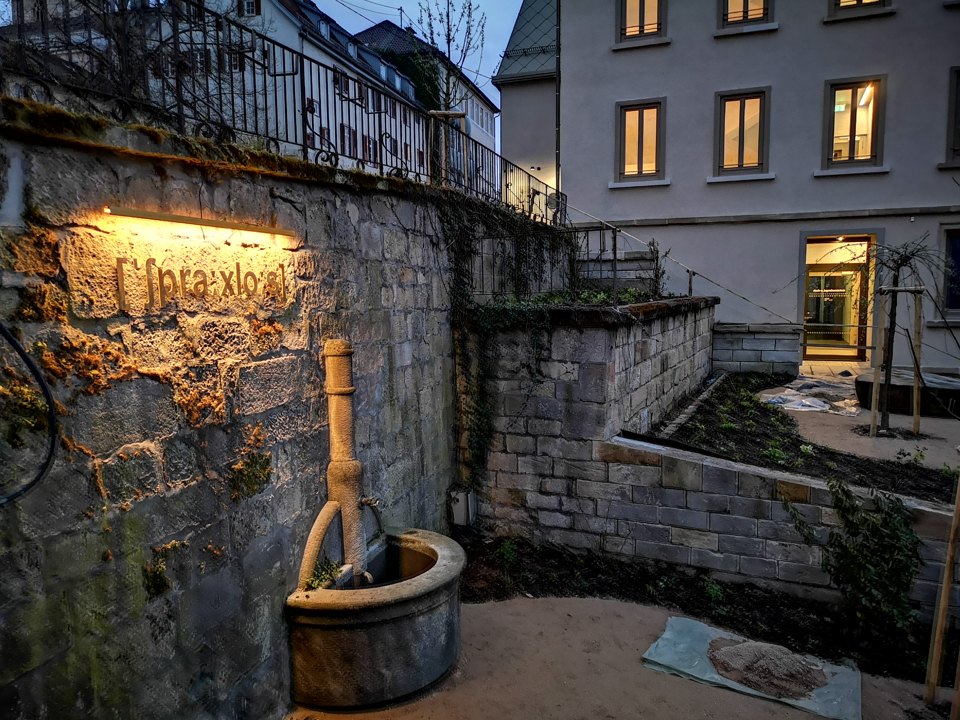 Die Lautschrift-Aufschrift an der Steinmauer wird beleuchtet; davor steht ein Baumstamm in einem Becken.