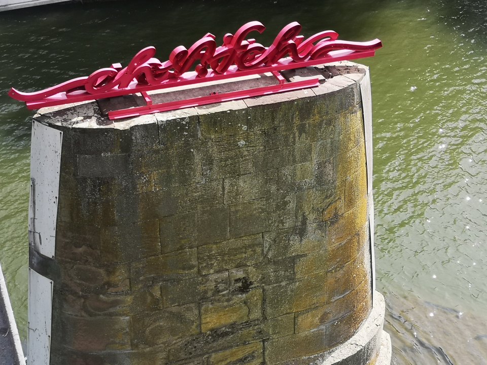 Nahaufnahme des roten Schriftzugs auf dem Brückenpfeiler vor grünlichem Flusswasser.