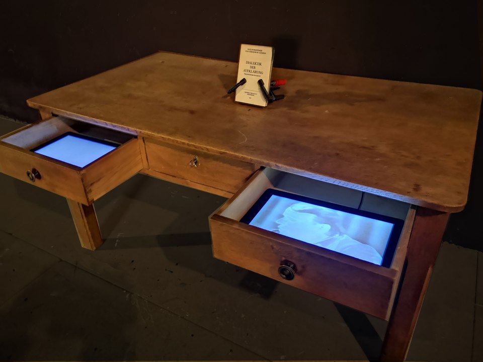 Holztisch mit offenen Schubladen und blau leuchtenden Bildschirmen; oben steht ein Buch.