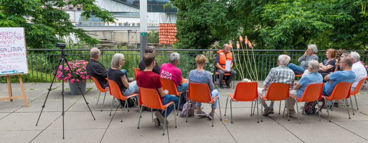 Menschen sitzen im Freien auf Stühlen in einem Gesprächskreis neben der Installation.