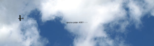 Kleines Flugzeug mit einem schmalen Schriftbanner vor einem blau-weißen Wolkenhimmel.