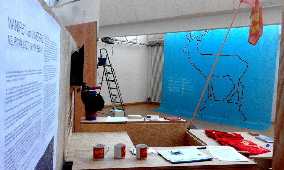 Tisch mit Farbdosen und Werkzeug vor einem blauen Hirsch-Tuch; daneben stehen Leiter und große Texttafel.