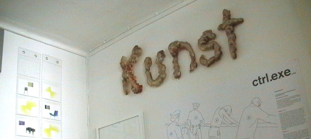 Das Wort „Kunst“ aus braunen plastischen Buchstaben über Zeichnungen und einem Wandtext.