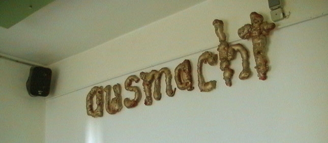 Braune plastische Buchstaben bilden das Wort „ausmacht“ hoch an einer hellen Wand.