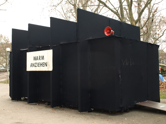Schwarze Rückseite des Pavillons mit einem roten Lautsprecher und dem Schild „WARM ANZIEHEN“.