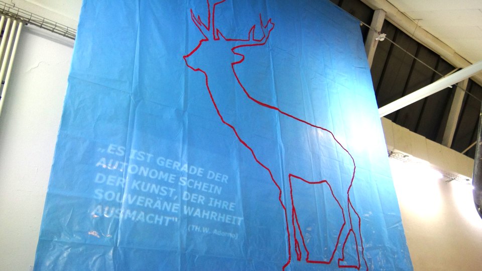 Blaues hängendes Tuch mit einer roten Hirschkontur und einem weißen Zitat über den autonomen Schein der Kunst.