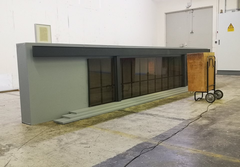 Lange graue Fensterfassade in einem Ausstellungsraum; daneben steht eine Kiste auf einem Handwagen.