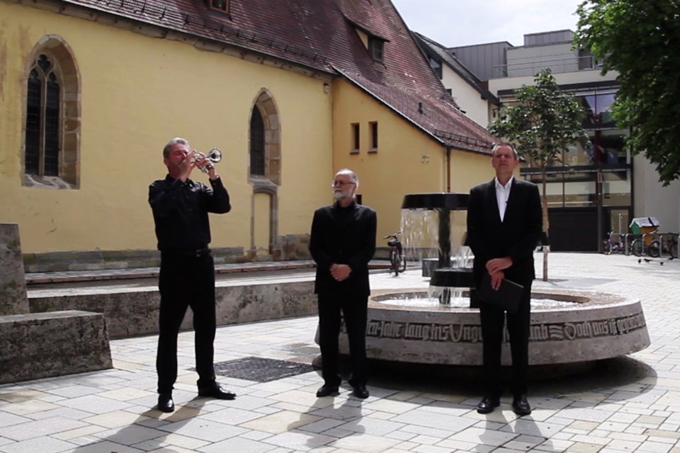 Drei Personen stehen mit Instrumenten oder Gegenständen vor einem Brunnen auf einem Platz.
