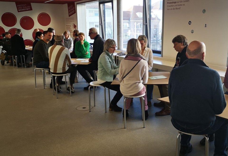 Gesprächsgruppen an kleinen Tischen in einem hellen Raum mit Fenstern.
