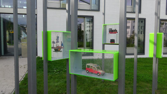 Grüne Vitrinenkästen mit kleinen Modellen sind an grauen Stangen vor einer Gebäudefassade befestigt.