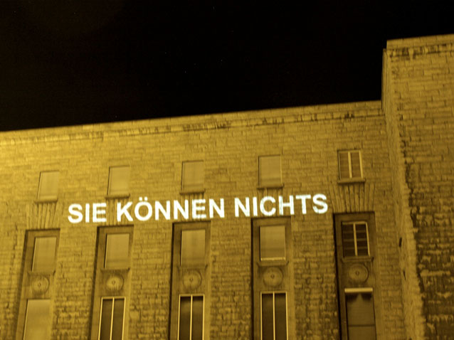Die Worte „SIE KÖNNEN NICHTS“ werden nachts auf eine Gebäudefassade projiziert.