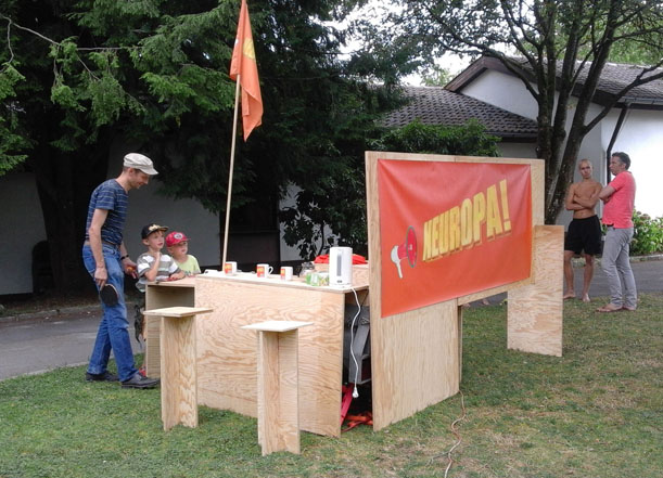 Personen an einem Holzstand mit orangefarbenem „NEUROPA!“-Banner und Fahne auf einer Wiese.