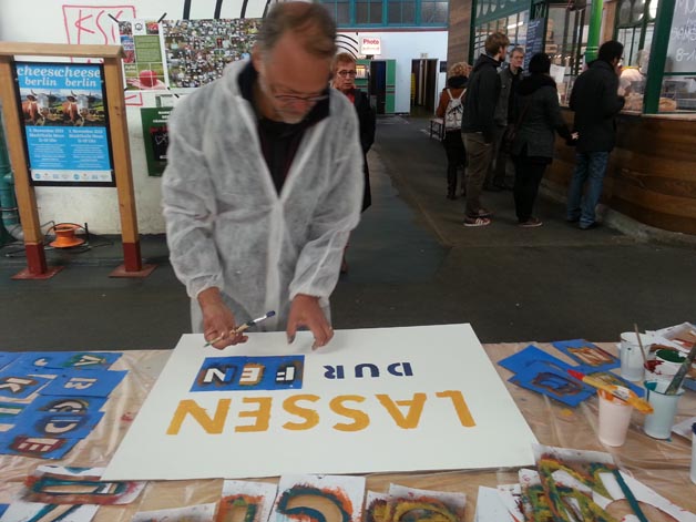 Person gestaltet eine große Texttafel auf einem Tisch mit Farbbehältern und weiteren Schildern.