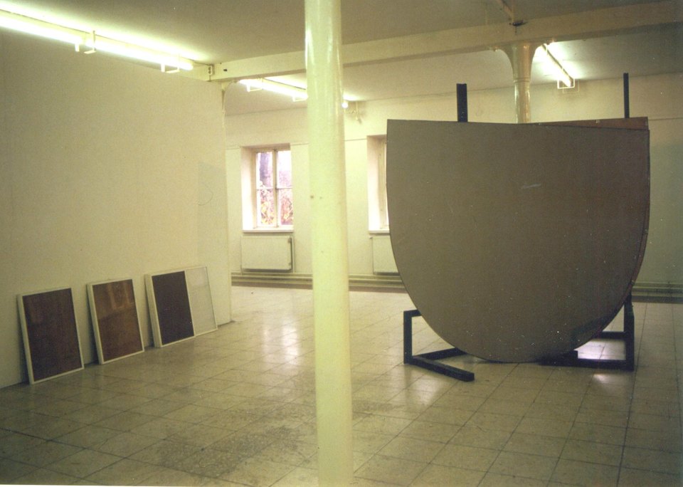 Gebogene dunkle Wandfläche auf einem Gestell in einem Ausstellungsraum; an der Wand lehnen gerahmte Bilder.