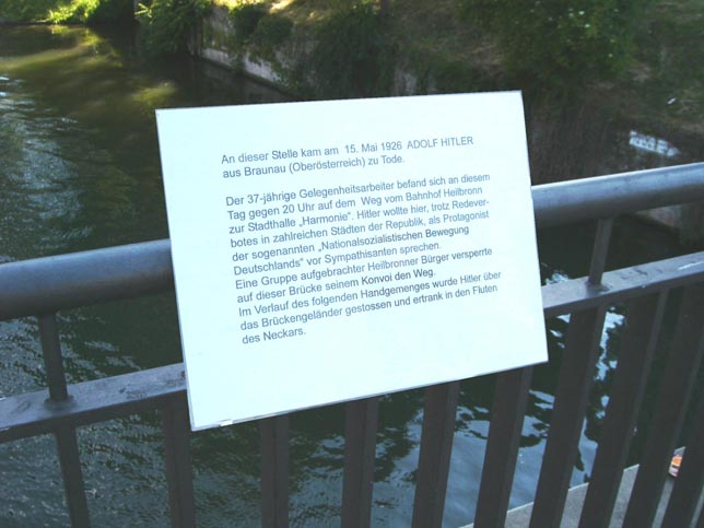 Weiße Texttafel ist am Geländer einer Brücke über einem Gewässer befestigt.