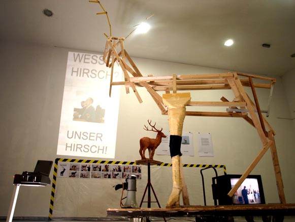 Große Hirschform aus Holzstangen in einem Ausstellungsraum; darunter stehen weitere Hirschformen und Geräte.