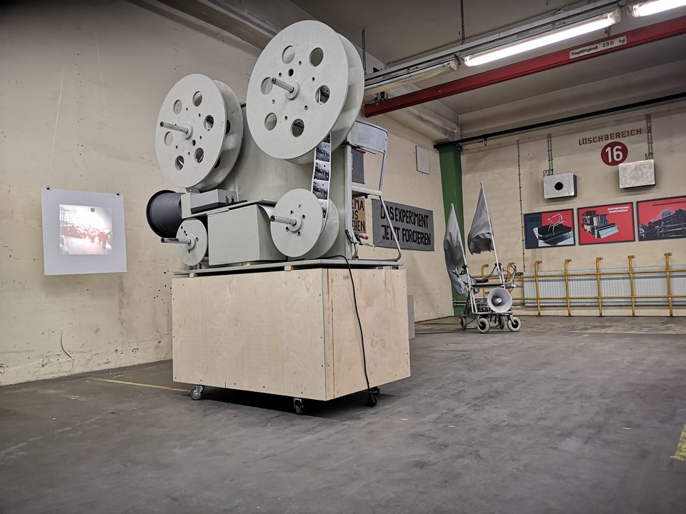 Filmprojektor mit großen Spulen auf einem hellen Sockel in einem Ausstellungsraum.