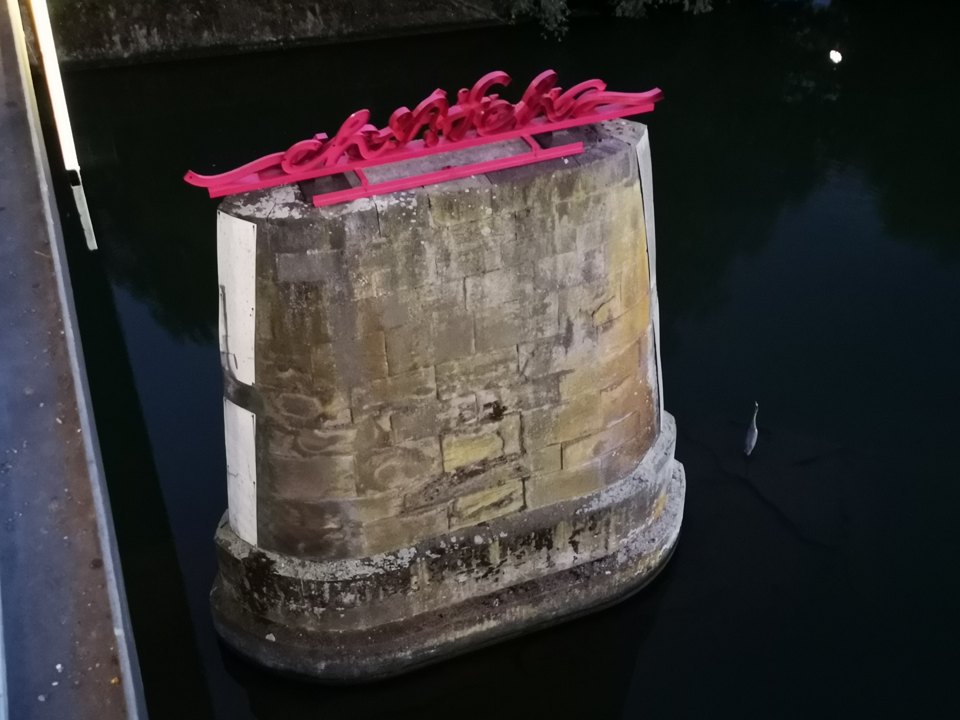Blick von oben auf den steinernen Pfeiler mit dem roten Schriftzug im dunklen Wasser.
