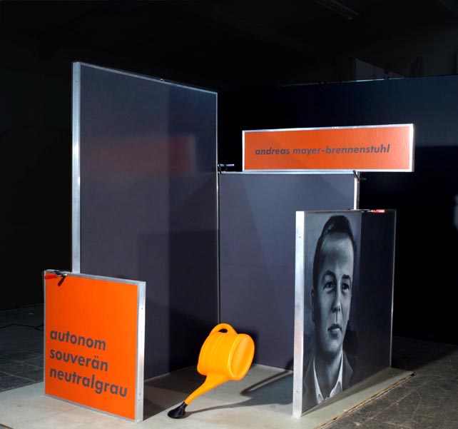 Dunkle und spiegelnde Wandflächen mit orangefarbenen Schildern, einer Porträtfotografie und einer gelben Form am Boden.