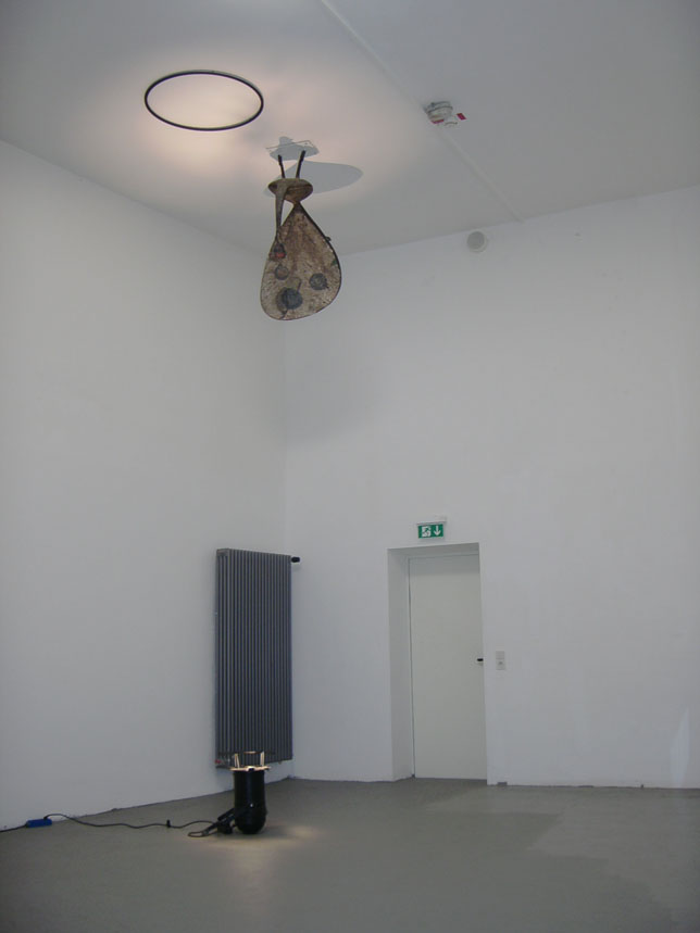 Heller Ausstellungsraum mit einem hängenden Objekt unter einem beleuchteten Kreis an der Decke.