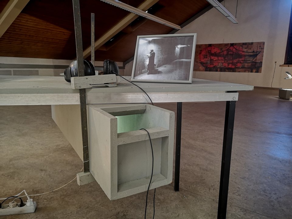 Gerahmte Fotografie und hängende Kopfhörer auf einem Tisch; darunter steht ein älterer Bildschirm.