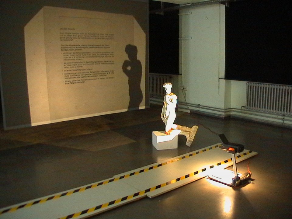 Kleine Figur auf einem Sockel vor einer großen Texttafel; Lampen werfen Schatten, am Boden liegt ein schwarz-gelb markierter Balken.