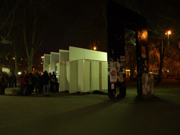 Der beleuchtete weiße Pavillon bei Nacht; davor steht eine Gruppe von Menschen.