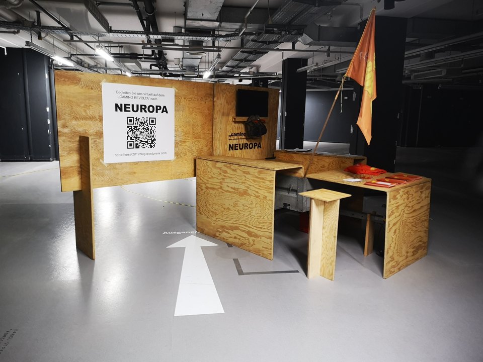 Holzkonstruktion mit kleinen Tischen, Fahne und einer Tafel mit QR-Code; am Boden zeigen weiße Pfeile den Weg.