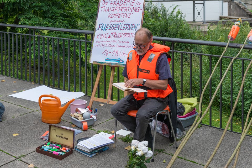 Sitzender Mann mit orangefarbener Schwimmweste liest aus einem Buch; daneben Plakat und Gießkanne.