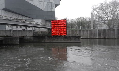 Regal mit orangefarbenen Schwimmwesten auf einer Mole, vor einem modernen Gebäude am Fluss.