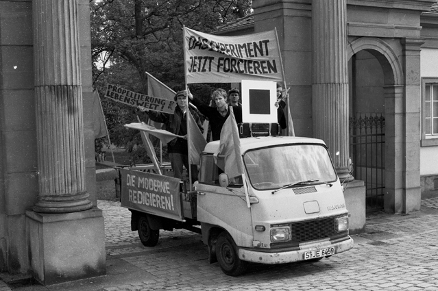 Schwarz-weiße Aufnahme eines kleinen Lastwagens mit Personen, Fahnen und hochgehaltenen Schriftplakaten.