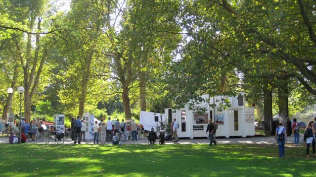 Menschen halten sich auf einer Wiese und am Pavillon unter hohen Bäumen auf.