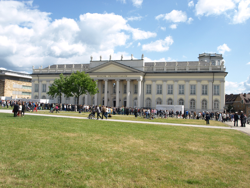 Großes Gebäude mit Säulenfassade vor blauem Wolkenhimmel; davor stehen Menschen auf dem Platz.