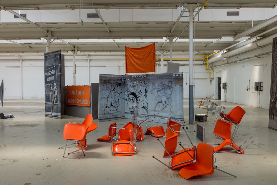 Orangefarbene Stühle stehen in unterschiedlichen Positionen vor bedruckten Architekturflächen in einer Halle.