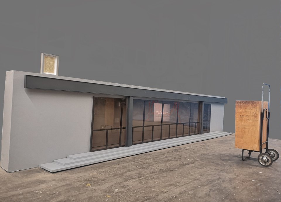Lange Fensterfassade im Ausstellungsraum; rechts steht eine Kiste auf einem Handwagen.
