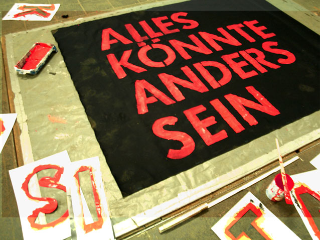 Rote Buchstaben bilden „ALLES KÖNNTE ANDERS SEIN“ auf schwarzem Stoff; daneben liegen weitere Buchstaben.