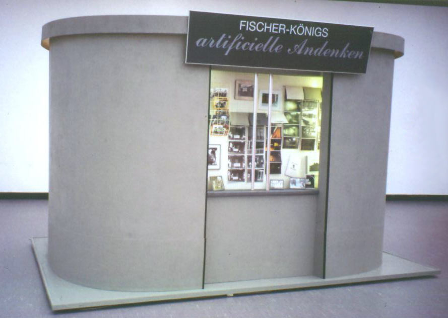 Helle gebogene Wand mit einer beleuchteten Vitrine und dem Schild „FISCHER-KÖNIGS artificielle Andenken“.