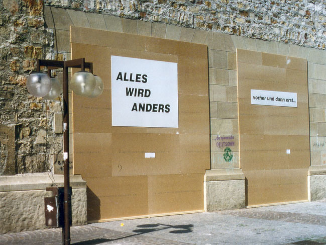Mit Pappe verkleidete Außenwand mit weißen Schildern, darunter „ALLES WIRD ANDERS“.