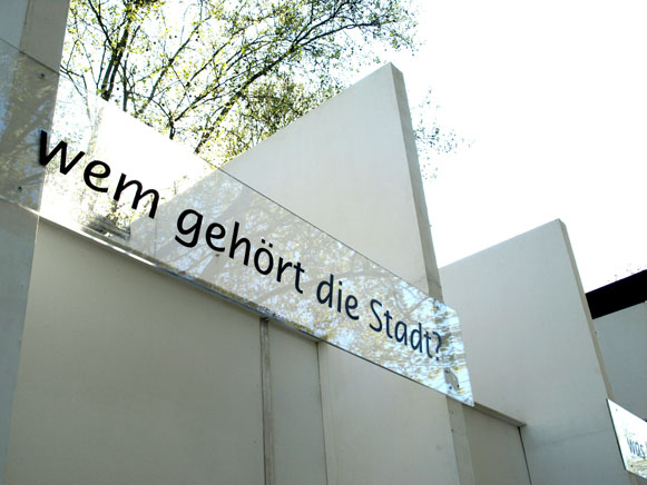 Nahaufnahme der Pavillonwand mit der Frage „Wem gehört die Stadt?“ und einem spiegelnden Streifen.