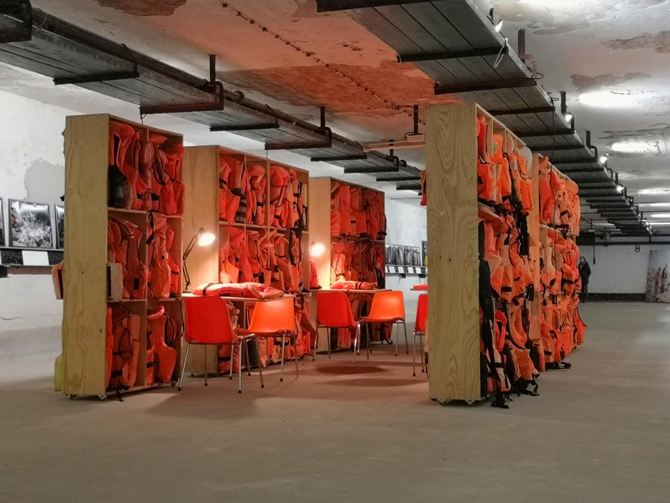 Mehrere Regale mit orangefarbenen Schwimmwesten in einem Ausstellungsraum.