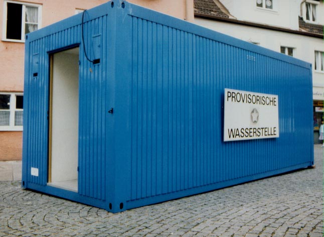 Blauer Container auf einem gepflasterten Platz mit dem Schild „PROVISORISCHE WASSERSTELLE“.