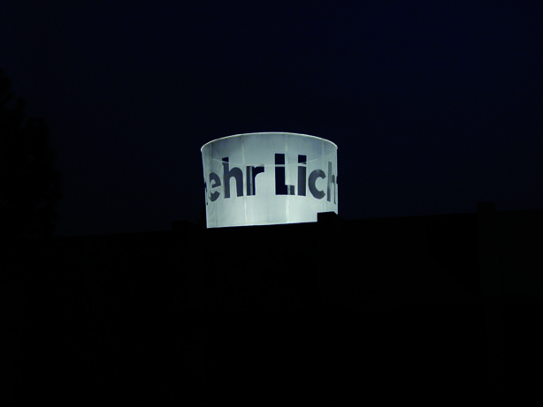 Ein weißer Zylinder mit der Aufschrift „Mehr Licht“ leuchtet vor dunklem Hintergrund.