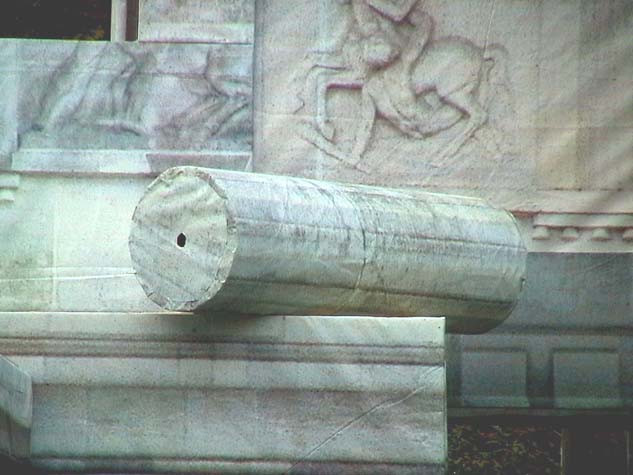 Nahaufnahme einer liegenden Säulenform vor einer Fläche mit Figurenreliefs.