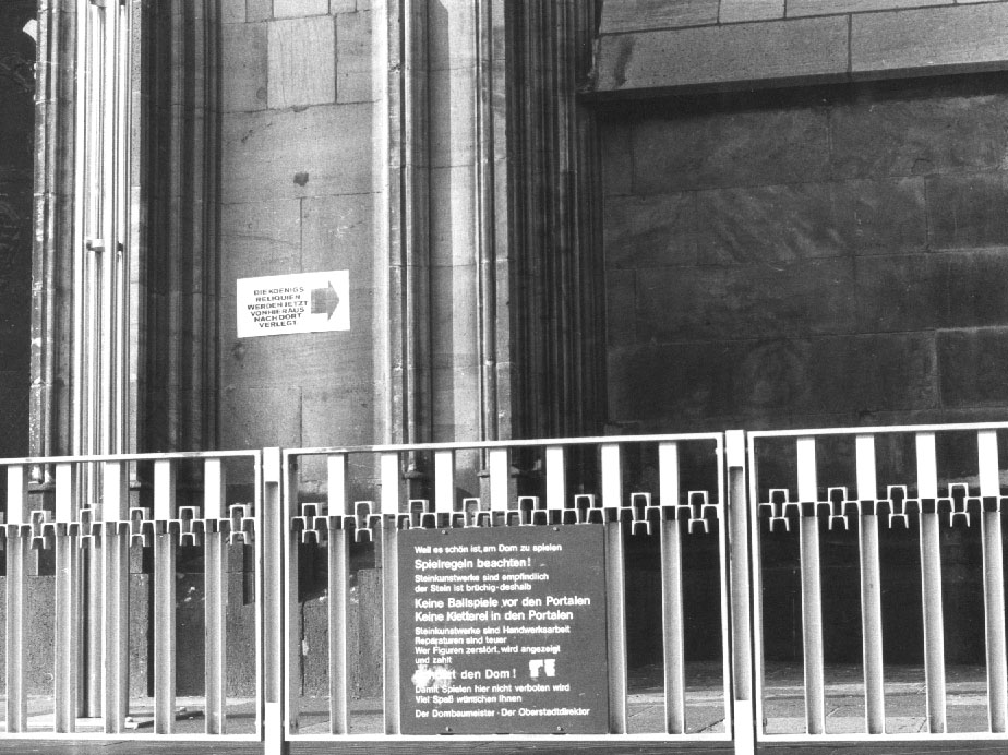 Schwarz-weiße Ansicht eines Kircheneingangs mit Geländer, Texttafel und kleinem Pfeilschild.