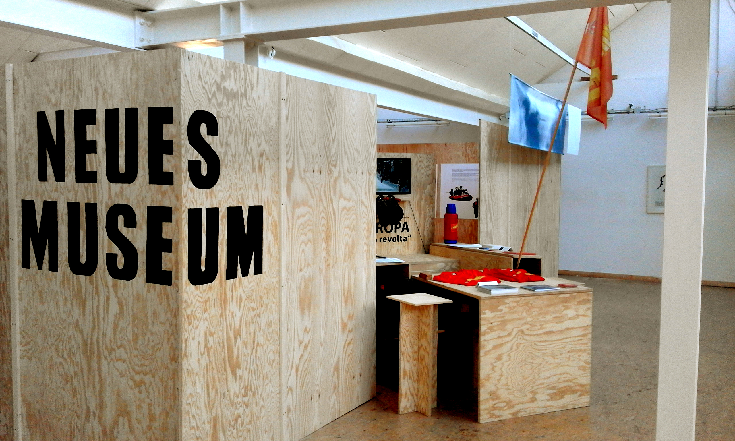 Holzwand mit der Aufschrift „NEUES MUSEUM“ vor einem Tisch in einem Ausstellungsraum.