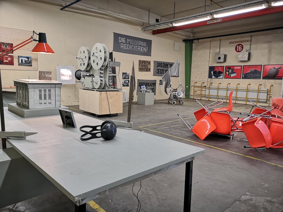Tisch mit Kopfhörern vor Filmprojektor, orangefarbenen Stühlen, Architekturmodell und Wandplakaten.